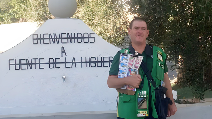 A ONCE lottery seller in a green vest holds a board of lottery tickets beside a white sign that reads ‘Bienvenidos a Fuente de la Higuera’.