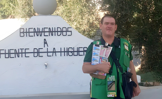 A ONCE lottery seller in a green vest holds a board of lottery tickets beside a white sign that reads ‘Bienvenidos a Fuente de la Higuera’.
