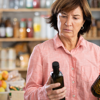 A woman in a pink shirt frowns as she compares two bottles of olive oil in a shop, with shelves of bottles, jars and fruit behind her