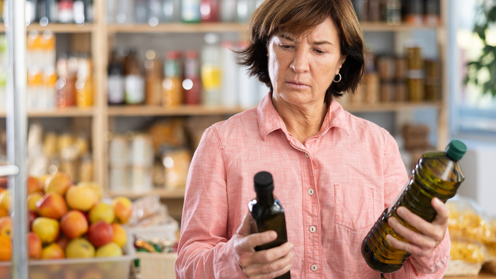 A woman in a pink shirt frowns as she compares two bottles of olive oil in a shop, with shelves of bottles, jars and fruit behind her