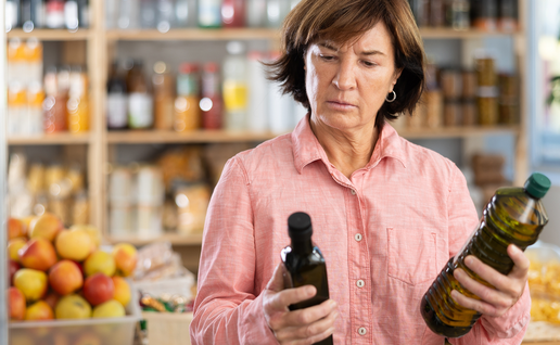 A woman in a pink shirt frowns as she compares two bottles of olive oil in a shop, with shelves of bottles, jars and fruit behind her