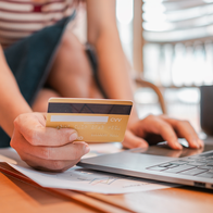 A person holding a gold bank card in one hand while typing on a laptop, with papers and a dark mug on the desk.