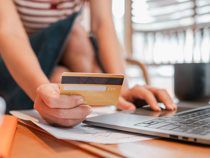 A person holding a gold bank card in one hand while typing on a laptop, with papers and a dark mug on the desk.