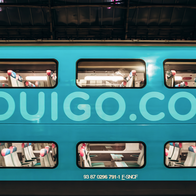 A turquoise double-decker train carriage with OUIGO.COM written along its side, standing under a station roof, with empty seats visible through the windows.