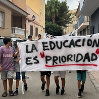 Children carry a hand-painted banner reading ‘La educación es prioridad’ down a residential street, with parked cars and a cloudy sky behind them.