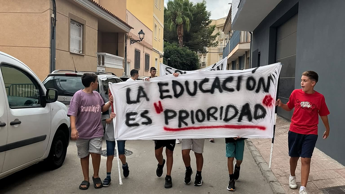 Children carry a hand-painted banner reading ‘La educación es prioridad’ down a residential street, with parked cars and a cloudy sky behind them.