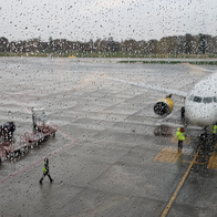 A passenger jet with yellow engines parked at a gate on a wet airport apron, seen through a rain-spotted window, with ground crew in hi-vis jackets and baggage trolleys nearby.