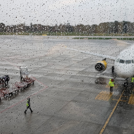 A passenger jet with yellow engines parked at a gate on a wet airport apron, seen through a rain-spotted window, with ground crew in hi-vis jackets and baggage trolleys nearby.
