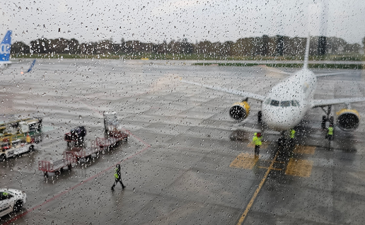 A passenger jet with yellow engines parked at a gate on a wet airport apron, seen through a rain-spotted window, with ground crew in hi-vis jackets and baggage trolleys nearby.