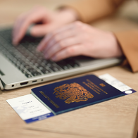 A blue British passport with a boarding pass tucked inside, on a wooden desk beside a laptop, with a person typing on the keyboard in the background.