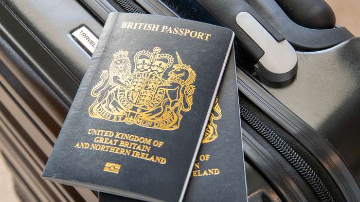 Two dark blue British passports with gold lettering resting on a black suitcase beside its handle.