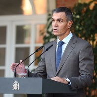 Pedro Sánchez speaking at a lectern with the Spanish coat of arms and two microphones, in front of a brick wall and a white arched window