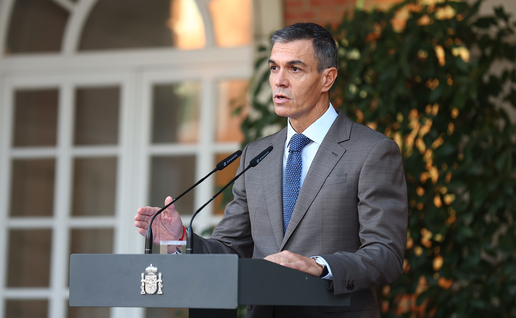 Pedro Sánchez speaking at a lectern with the Spanish coat of arms and two microphones, in front of a brick wall and a white arched window