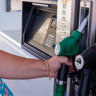 A man’s hand holds a green petrol nozzle at a fuel pump, next to a black nozzle marked Gasoil A.