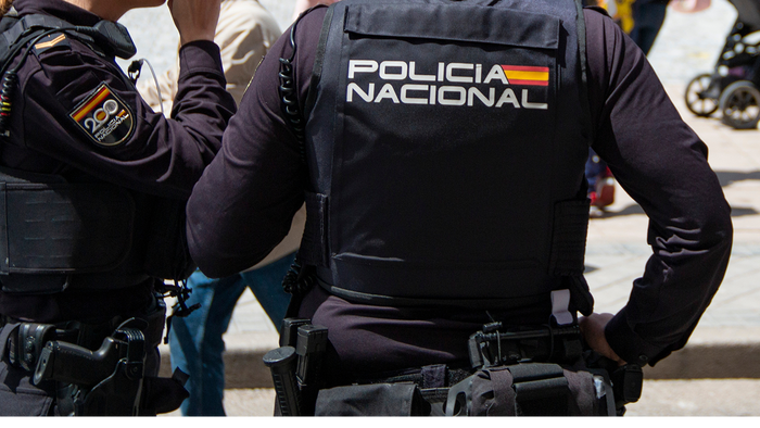 Two Policía Nacional officers seen from behind on a sunny street, one in a dark vest marked Policía Nacional.
