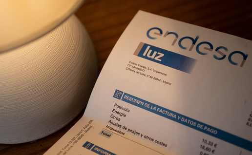 An Endesa electricity bill showing a summary of charges, on a wooden table next to a lit lamp.