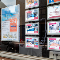 Estate agent's shop window with rows of illuminated panels showing homes for sale, alongside a poster for a new-build development.