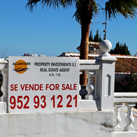 A white “Se vende, for sale” estate agent sign with a red phone number on a whitewashed garden wall, with palm trees, a terracotta tiled roof and blue sky behind.