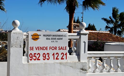 A white “Se vende, for sale” estate agent sign with a red phone number on a whitewashed garden wall, with palm trees, a terracotta tiled roof and blue sky behind.