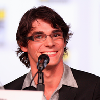 RJ Mitte, a young man with dark wavy hair and glasses, smiling into a microphone at a convention panel, wearing a dark blazer over a grey shirt.