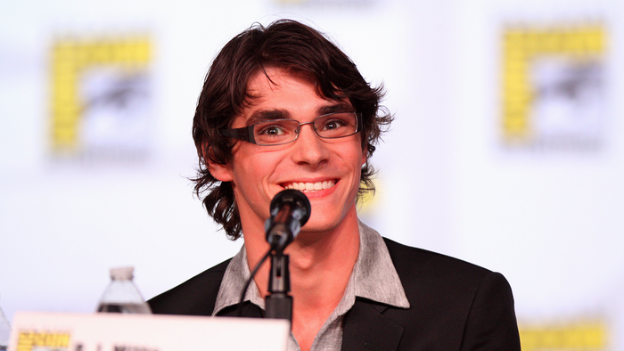 RJ Mitte, a young man with dark wavy hair and glasses, smiling into a microphone at a convention panel, wearing a dark blazer over a grey shirt.