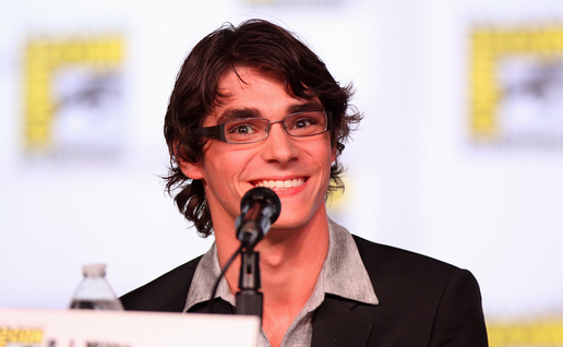 RJ Mitte, a young man with dark wavy hair and glasses, smiling into a microphone at a convention panel, wearing a dark blazer over a grey shirt.
