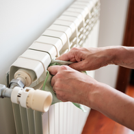 Two hands wiping the top of a white panel radiator with a green cloth, with the thermostatic valve and pipe in the foreground and a window behind.