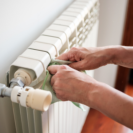 Two hands wiping the top of a white panel radiator with a green cloth, with the thermostatic valve and pipe in the foreground and a window behind.
