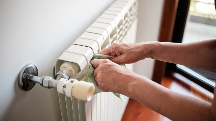 Two hands wiping the top of a white panel radiator with a green cloth, with the thermostatic valve and pipe in the foreground and a window behind.