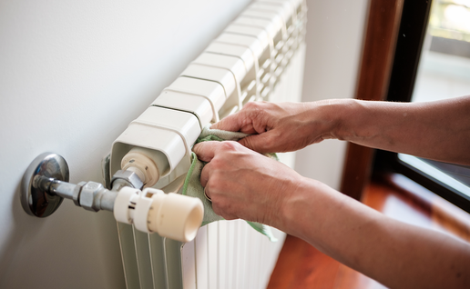 Two hands wiping the top of a white panel radiator with a green cloth, with the thermostatic valve and pipe in the foreground and a window behind.