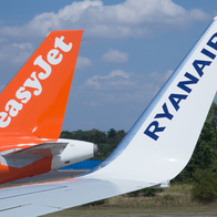 Tail fins of an orange easyJet plane and a white Ryanair plane side by side under a blue sky.