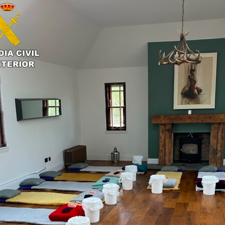 Large room with wooden floors and high ceiling, lined with rows of mattresses, pillows, blankets and white plastic buckets; Guardia Civil (Spanish Civil Guard) logo in the top-left corner.