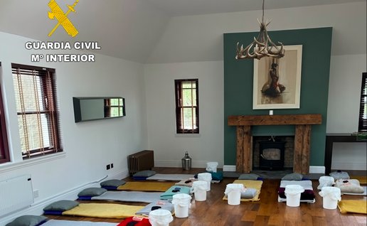Large room with wooden floors and high ceiling, lined with rows of mattresses, pillows, blankets and white plastic buckets; Guardia Civil (Spanish Civil Guard) logo in the top-left corner.
