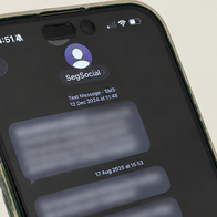 A hand holding a smartphone that shows a text message thread from a sender named SegSocial, with the message text blurred out.