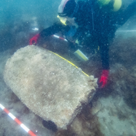 Diver in a black wetsuit and red gloves leaning over a large, rounded, barrel-shaped object covered in marine growth on the seabed, with a red-and-white scale bar and measuring tapes laid around it.