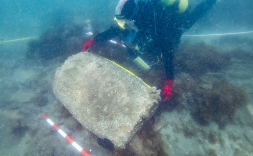 Diver in a black wetsuit and red gloves leaning over a large, rounded, barrel-shaped object covered in marine growth on the seabed, with a red-and-white scale bar and measuring tapes laid around it.