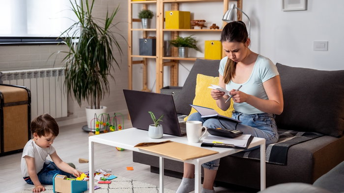 Mother reviewing household bills and paperwork at home while her young child plays nearby.