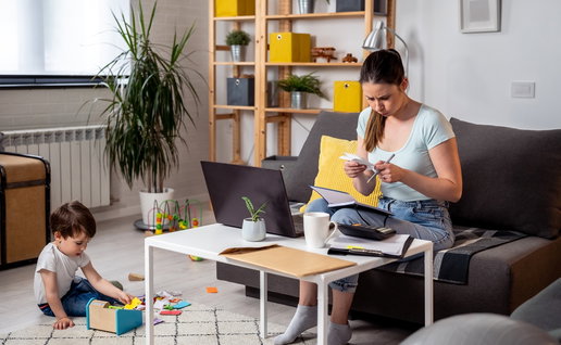 Mother reviewing household bills and paperwork at home while her young child plays nearby.