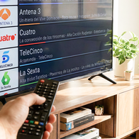 A hand points a remote control at a flat-screen television showing a list of Spanish channels, including La 1, Antena 3, Cuatro, Telecinco and La Sexta, in a bright living room with plants.