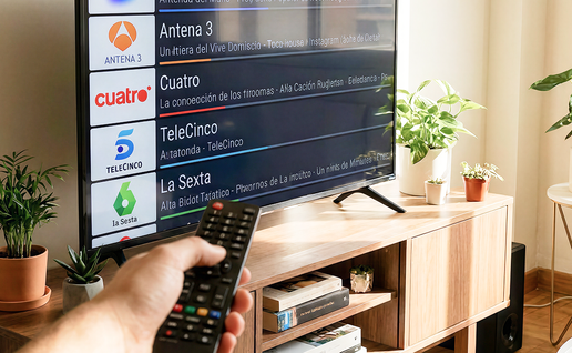 A hand points a remote control at a flat-screen television showing a list of Spanish channels, including La 1, Antena 3, Cuatro, Telecinco and La Sexta, in a bright living room with plants.