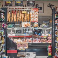 A supermarket aisle with shelves of oils and jars on both sides, leading to a deli counter with legs of ham hanging above it and a member of staff wearing blue gloves.