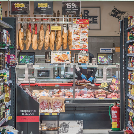 A supermarket aisle with shelves of oils and jars on both sides, leading to a deli counter with legs of ham hanging above it and a member of staff wearing blue gloves.