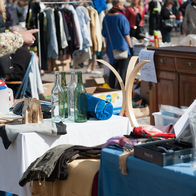 Shoppers browse crowded outdoor flea market stalls, with glass bottles, vases and clothes on tables, a rack of clothing and a wooden chest of drawers for sale.