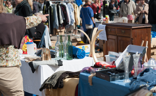 Shoppers browse crowded outdoor flea market stalls, with glass bottles, vases and clothes on tables, a rack of clothing and a wooden chest of drawers for sale.