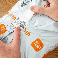 Two hands holding a grey plastic parcel printed with orange Temu logos, with a shipping label and QR code on the front, on a wooden table.