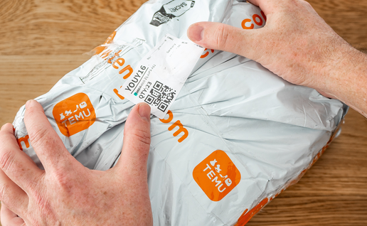 Two hands holding a grey plastic parcel printed with orange Temu logos, with a shipping label and QR code on the front, on a wooden table.