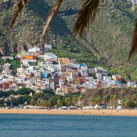 Colourful houses climbing a green mountainside above a sandy beach and calm blue bay, seen through palm fronds.