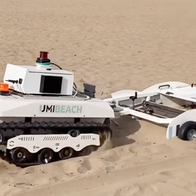 A white tracked robot with “UMIBEACH” on its side, towing a wheeled sieving trailer across a sandy beach.