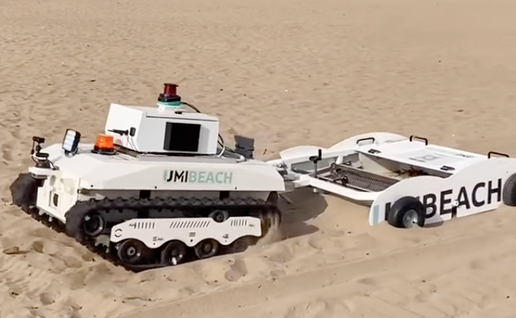 A white tracked robot with “UMIBEACH” on its side, towing a wheeled sieving trailer across a sandy beach.