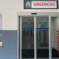 Entrance to a hospital emergency department with a green and white Junta de Andalucía “Urgencias” sign above sliding glass doors.